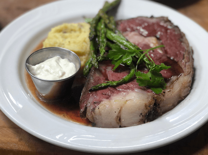 A plated meal featuring a juicy roast beef with asparagus, creamy mashed potatoes, and a small cup of sauce.