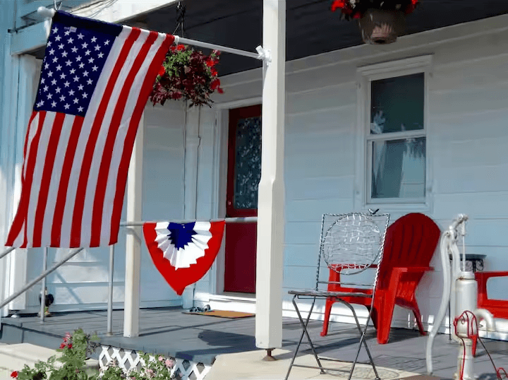 A front porch decorated with an American flag, patriotic bunting, and red chairs.